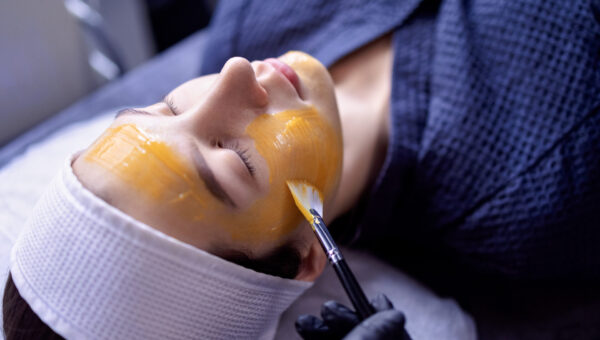 A young woman does a cosmetic procedure at a beauty center or clinic. The cosmetologist applies a rejuvenating orange mask to the client face. Facelift and skin care at the spa.